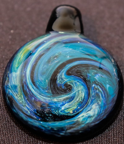 A handcrafted glass pendant with intricate swirling patterns resembling a blue and green galaxy or whirlpool, set against a dark background. The design includes shimmering elements that add a subtle sparkle.