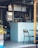 A modern kitchen setup with a small bar area. There are appliances like an oven and a display of jars, bottles, and fruits on the counter. The decor features white tiles with red grout and simple chairs and tables.
