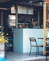 A modern kitchen setup with a small bar area. There are appliances like an oven and a display of jars, bottles, and fruits on the counter. The decor features white tiles with red grout and simple chairs and tables.
