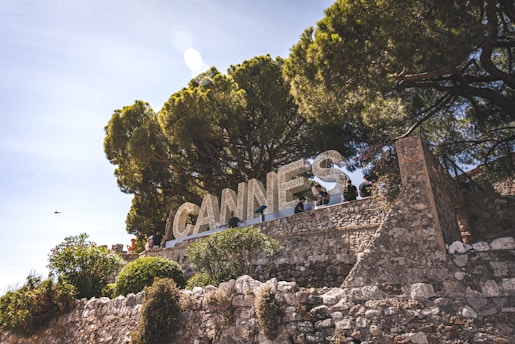 Large white letters spelling 'CANNES' are mounted on a stone terrace, partially surrounded by lush green trees. Several people are standing or walking near the letters, enjoying the sunny day.