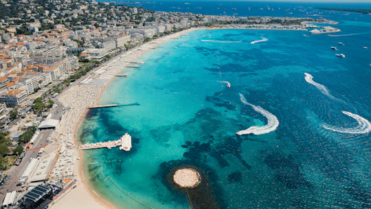 A bustling coastline with clear turquoise waters, dotted with boats and marked by the wakes of watercraft. The sandy beach is lined with rows of sunbathers under parasols, and the urban area with numerous buildings stretches into the distance. The scene captures a lively, summery beach locale with vibrant coastal activity.