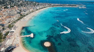 A bustling coastline with clear turquoise waters, dotted with boats and marked by the wakes of watercraft. The sandy beach is lined with rows of sunbathers under parasols, and the urban area with numerous buildings stretches into the distance. The scene captures a lively, summery beach locale with vibrant coastal activity.
