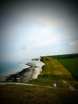 A vast coastline with white chalk cliffs stretching alongside a grassy hill that slopes down towards the sea. The ocean is partially visible on the left side, with its waters gently meeting the pebbled shoreline. Several people are walking and standing on the grass, enjoying the scenic view.