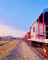 A train with red and white carriages travels along railway tracks through a rural landscape. The scene is set against a clear, blue sky with a hint of gradient as the sun sets or rises. Green hills and patches of trees are visible in the background.