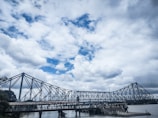 A large steel bridge spans across a river, under an expansive sky filled with fluffy white and grey clouds. Below the bridge, a small dock area with people can be seen near the water's edge. The structure conveys a sense of engineering prowess against a dramatic cloudy backdrop.