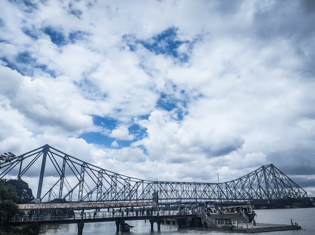 A large steel bridge spans across a river, under an expansive sky filled with fluffy white and grey clouds. Below the bridge, a small dock area with people can be seen near the water's edge. The structure conveys a sense of engineering prowess against a dramatic cloudy backdrop.