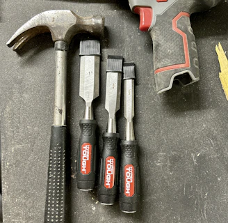A collection of tools is arranged on a dark surface. An electric drill with the brand 'Hyper Tough' is visible alongside a claw hammer with a black handle. Three chisels of varying sizes, also branded 'Hyper Tough,' are aligned next to the hammer.