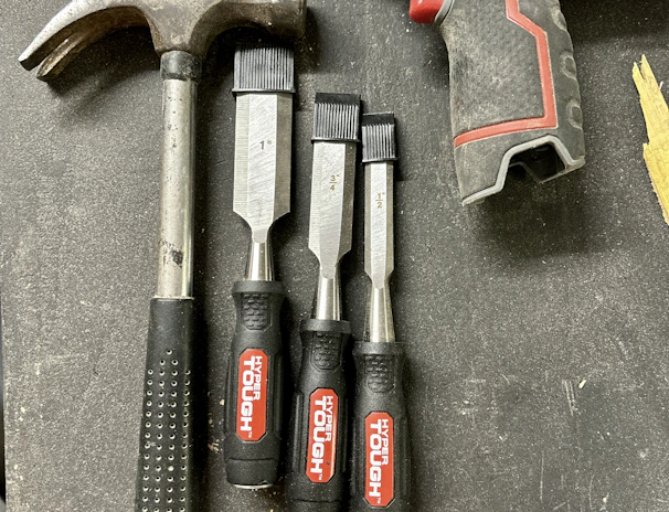 A collection of tools is arranged on a dark surface. An electric drill with the brand 'Hyper Tough' is visible alongside a claw hammer with a black handle. Three chisels of varying sizes, also branded 'Hyper Tough,' are aligned next to the hammer.