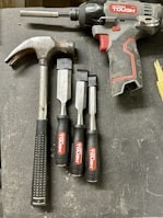 A collection of tools is arranged on a dark surface. An electric drill with the brand 'Hyper Tough' is visible alongside a claw hammer with a black handle. Three chisels of varying sizes, also branded 'Hyper Tough,' are aligned next to the hammer.