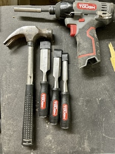 A collection of tools is arranged on a dark surface. An electric drill with the brand 'Hyper Tough' is visible alongside a claw hammer with a black handle. Three chisels of varying sizes, also branded 'Hyper Tough,' are aligned next to the hammer.