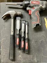 A collection of tools is arranged on a dark surface. An electric drill with the brand 'Hyper Tough' is visible alongside a claw hammer with a black handle. Three chisels of varying sizes, also branded 'Hyper Tough,' are aligned next to the hammer.