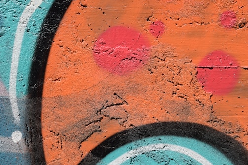 Vibrant graffiti art displaying dynamic curves and shapes, with a textured wall as the canvas. The artwork features prominent splashes of orange and teal, intersected by bold black and white lines and circles in varying shades of bright pink.