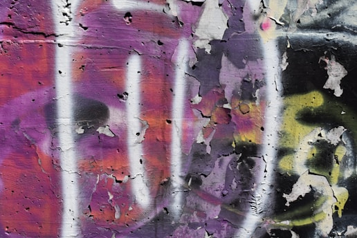 Peeling paint on a wall reveals colorful graffiti with a mix of purple, pink, black, white, and yellow hues. The surface appears weathered and textured with cracks and chipped paint.