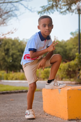 A young boy poses confidently outdoors, with one knee bent on a yellow block and giving a peace sign. He is wearing a light blue polo shirt with red accents and beige shorts. The background is a park-like setting with trees and greenery.