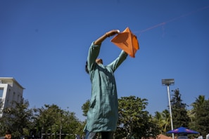 A person is flying an orange kite against a clear blue sky. The individual is wearing a teal kurta and standing in an open area, with trees and a stadium light visible in the background.