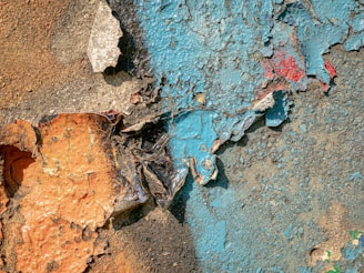 Chipped and peeling paint on a rough, textured surface with layers of rusty orange, blue, and bits of red color.