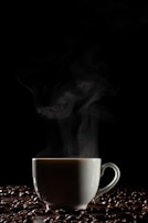 A white ceramic cup is filled with steaming hot coffee, surrounded by scattered coffee beans on a dark surface. The rising steam creates a contrasting effect against the black background, highlighting the warmth and aroma of the beverage.