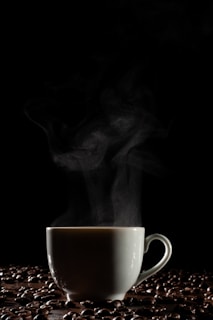 A white ceramic cup is filled with steaming hot coffee, surrounded by scattered coffee beans on a dark surface. The rising steam creates a contrasting effect against the black background, highlighting the warmth and aroma of the beverage.