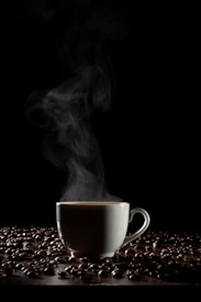 A steaming cup of coffee is enveloped by rising vapor against a dark background. The cup is surrounded by scattered coffee beans on a wooden surface, casting subtle shadows. The atmosphere conveys a sense of warmth and coziness.