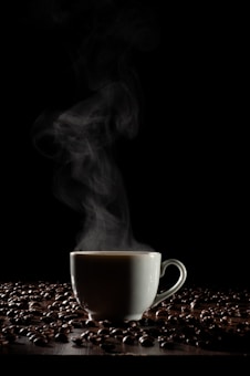 A steaming cup of coffee is enveloped by rising vapor against a dark background. The cup is surrounded by scattered coffee beans on a wooden surface, casting subtle shadows. The atmosphere conveys a sense of warmth and coziness.