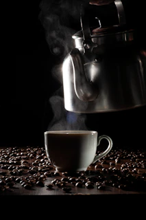 A metal kettle is pouring hot liquid, likely coffee, into a white cup surrounded by scattered coffee beans. Steam rises from the cup, creating a warm and inviting atmosphere against a dark background.
