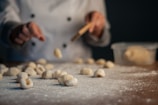 A person wearing a chef's coat is shaping gnocchi on a floured surface. The hands are in the process of working with the dough, and several pieces of gnocchi are spread out. The background is slightly blurred, adding depth to the scene.