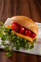 A fresh sandwich placed on a cloth napkin, featuring a crispy bread roll stuffed with slices of cheese, vibrant red cherry tomatoes, and a generous amount of green leafy lettuce. The background consists of a wooden surface which adds a rustic feel to the setup.