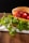 A fresh sandwich displayed on a wooden surface, featuring ripe red tomatoes and vibrant green leaves nestled between slices of bread. The bread appears soft and lightly toasted, and there's a thin slice of cheese visible underneath the tomatoes. A white and green cloth is spread underneath, adding a rustic touch to the presentation.