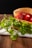 A fresh sandwich displayed on a wooden surface, featuring ripe red tomatoes and vibrant green leaves nestled between slices of bread. The bread appears soft and lightly toasted, and there's a thin slice of cheese visible underneath the tomatoes. A white and green cloth is spread underneath, adding a rustic touch to the presentation.