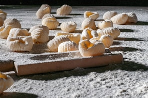 Handmade pasta pieces are scattered across a flour-dusted surface, with a rolling pin visible in the foreground. The textured pasta shapes are exposed to natural light, highlighting their ridged pattern and creating shadows on the surface.