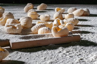 Handmade pasta pieces are scattered across a flour-dusted surface, with a rolling pin visible in the foreground. The textured pasta shapes are exposed to natural light, highlighting their ridged pattern and creating shadows on the surface.
