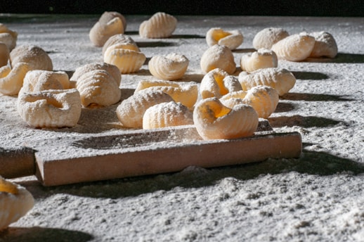 Handmade pasta pieces are scattered across a flour-dusted surface, with a rolling pin visible in the foreground. The textured pasta shapes are exposed to natural light, highlighting their ridged pattern and creating shadows on the surface.