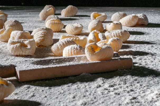 Handmade pasta pieces are scattered across a flour-dusted surface, with a rolling pin visible in the foreground. The textured pasta shapes are exposed to natural light, highlighting their ridged pattern and creating shadows on the surface.