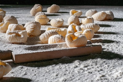 Handmade pasta pieces are scattered across a flour-dusted surface, with a rolling pin visible in the foreground. The textured pasta shapes are exposed to natural light, highlighting their ridged pattern and creating shadows on the surface.