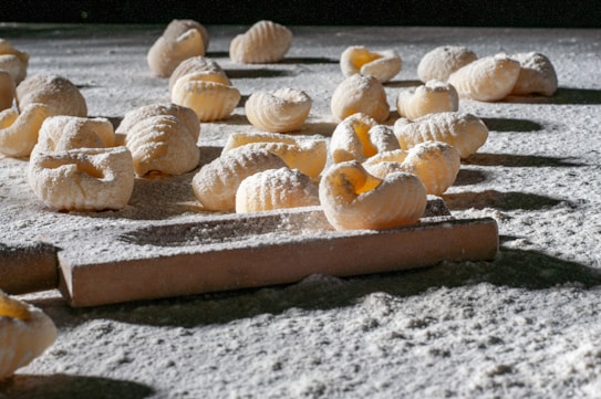 Handmade pasta pieces are scattered across a flour-dusted surface, with a rolling pin visible in the foreground. The textured pasta shapes are exposed to natural light, highlighting their ridged pattern and creating shadows on the surface.
