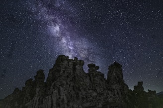 A starry night sky with a prominent Milky Way galaxy is visible above unique rock formations. The sky is filled with countless stars, and the galaxy's bright core and swirling patterns create a captivating display.
