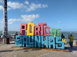 A colorful, large sign with the words 'PORTO DE GALINHAS' stands prominently on a paved promenade. The letters are a mix of red, yellow, blue, and green colors. In the background, the ocean is visible, along with a clear blue sky scattered with a few clouds. People are casually standing around the sign, and their expressions suggest a relaxed, leisurely atmosphere.