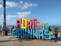 A colorful, large sign with the words 'PORTO DE GALINHAS' stands prominently on a paved promenade. The letters are a mix of red, yellow, blue, and green colors. In the background, the ocean is visible, along with a clear blue sky scattered with a few clouds. People are casually standing around the sign, and their expressions suggest a relaxed, leisurely atmosphere.