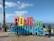 A colorful, large sign with the words 'PORTO DE GALINHAS' stands prominently on a paved promenade. The letters are a mix of red, yellow, blue, and green colors. In the background, the ocean is visible, along with a clear blue sky scattered with a few clouds. People are casually standing around the sign, and their expressions suggest a relaxed, leisurely atmosphere.