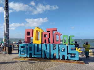 A colorful, large sign with the words 'PORTO DE GALINHAS' stands prominently on a paved promenade. The letters are a mix of red, yellow, blue, and green colors. In the background, the ocean is visible, along with a clear blue sky scattered with a few clouds. People are casually standing around the sign, and their expressions suggest a relaxed, leisurely atmosphere.