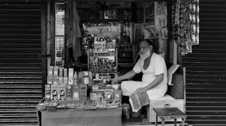 An elderly man with a long beard sits inside a small, densely packed shop. The shop sells various personal care items, including shampoo, soap, and other toiletries. The man is wearing a simple, light-colored outfit and appears to be waiting for customers. The shop is surrounded by closed shutters on either side.