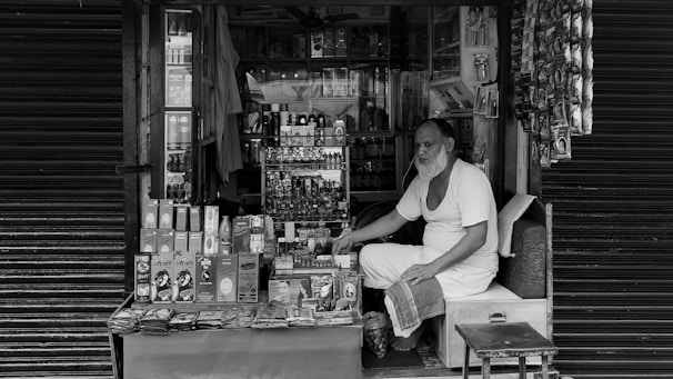 An elderly man with a long beard sits inside a small, densely packed shop. The shop sells various personal care items, including shampoo, soap, and other toiletries. The man is wearing a simple, light-colored outfit and appears to be waiting for customers. The shop is surrounded by closed shutters on either side.