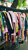 A variety of colorful clothes hang on black hangers outside on a wheeled cart. The collection consists of shirts, dresses, and tops in various colors and patterns, arranged closely together. Above them hangs a tree branch with green leaves visible in the background.