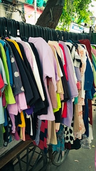 A variety of colorful clothes hang on black hangers outside on a wheeled cart. The collection consists of shirts, dresses, and tops in various colors and patterns, arranged closely together. Above them hangs a tree branch with green leaves visible in the background.