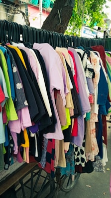 A variety of colorful clothes hang on black hangers outside on a wheeled cart. The collection consists of shirts, dresses, and tops in various colors and patterns, arranged closely together. Above them hangs a tree branch with green leaves visible in the background.