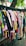 A variety of colorful clothes hang on black hangers outside on a wheeled cart. The collection consists of shirts, dresses, and tops in various colors and patterns, arranged closely together. Above them hangs a tree branch with green leaves visible in the background.
