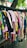 A variety of colorful clothes hang on black hangers outside on a wheeled cart. The collection consists of shirts, dresses, and tops in various colors and patterns, arranged closely together. Above them hangs a tree branch with green leaves visible in the background.