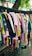 A variety of colorful clothes hang on black hangers outside on a wheeled cart. The collection consists of shirts, dresses, and tops in various colors and patterns, arranged closely together. Above them hangs a tree branch with green leaves visible in the background.