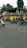 An urban street scene featuring several people riding scooters and motorcycles. Auto rickshaws are parked by the side, and there are buildings in the background with visible signage in a local script. The environment is bustling, with greenery from trees lining the street.