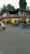 An urban street scene featuring several people riding scooters and motorcycles. Auto rickshaws are parked by the side, and there are buildings in the background with visible signage in a local script. The environment is bustling, with greenery from trees lining the street.